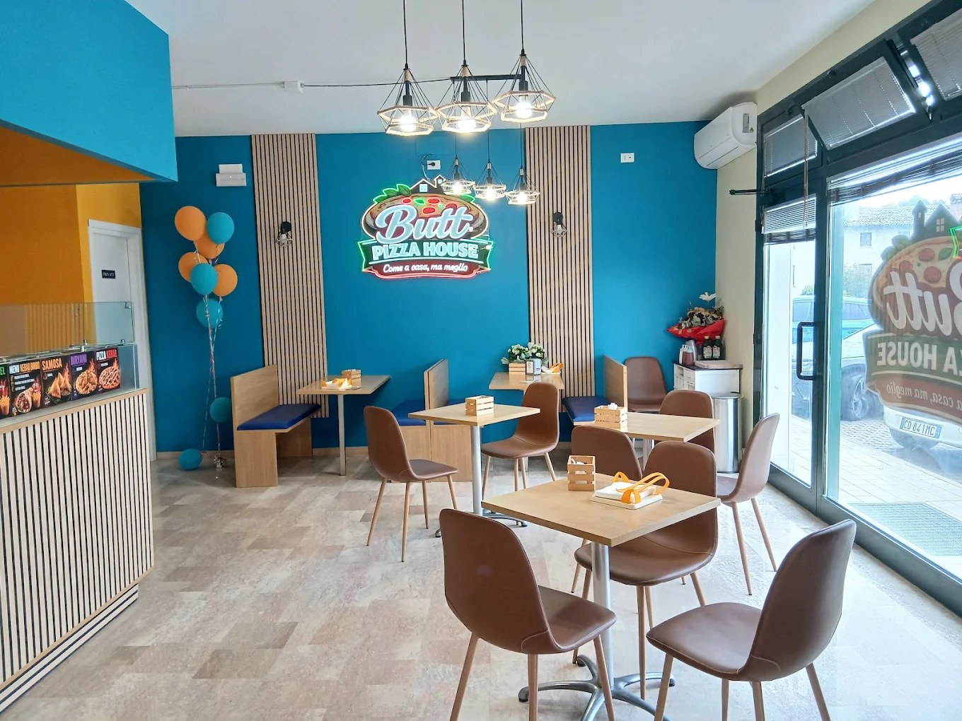 Sala di Butt Pizza House con insegna luminosa «Come a casa, ma meglio», pareti blu, doghe in legno e sedie marroni