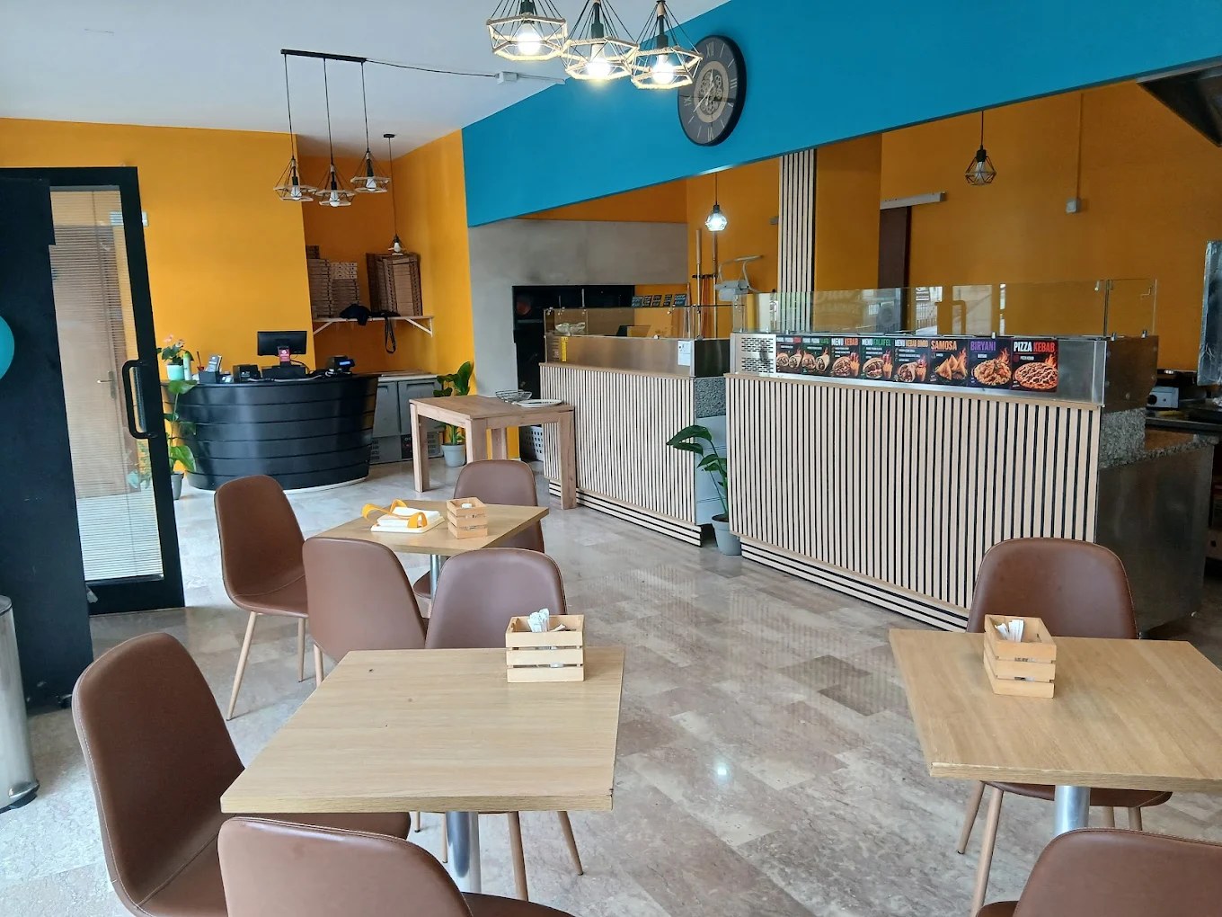 Sala con pareti gialle e blu, tavoli in legno, sedie marroni e bancone con pannello Samosa, Biryani, Pizza Kebab